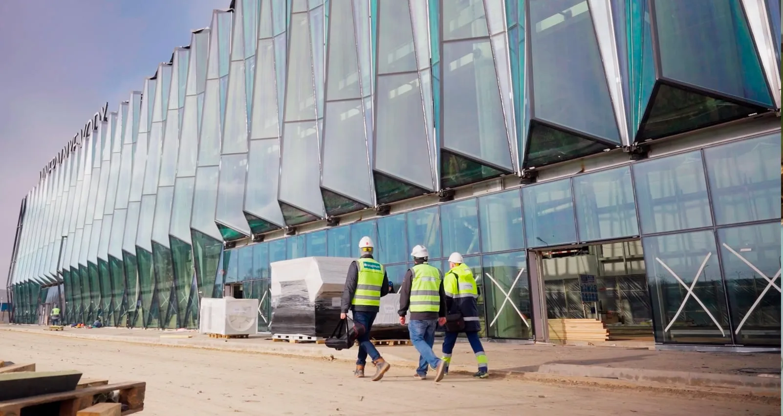 Three engineers in safety vests and helmets walk along the glass facade of the airport terminal under construction.