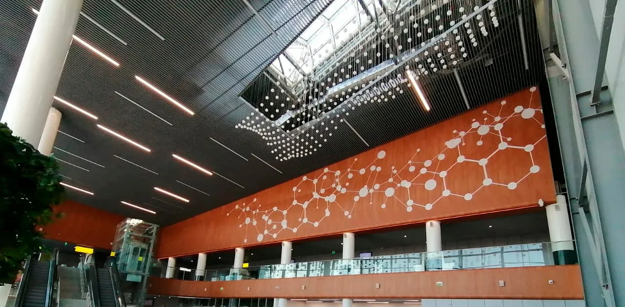 The hall of the airport terminal with a high ceiling, a skylight, escalators and a wall panel with a pattern of connected white dots.