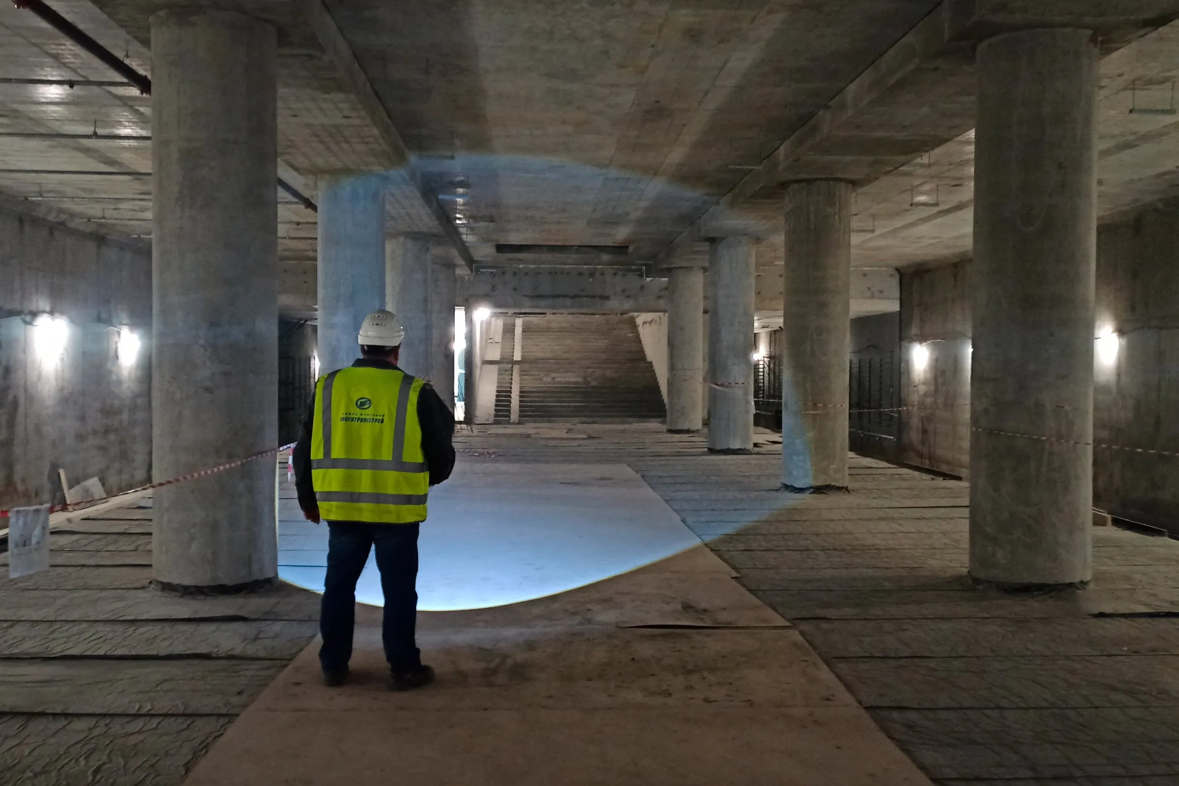 A worker in a safety vest and helmet stands in a concrete metro station hall under construction, with a flight of stairs ahead.
